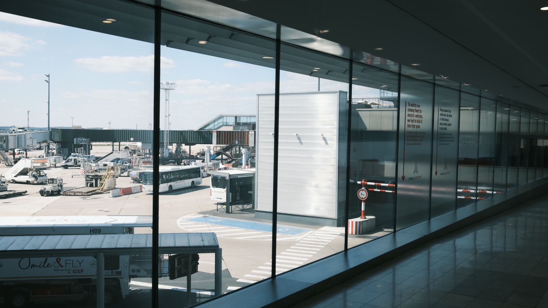 Wide view of an airport terminal hallway and tarmac, showcasing airplanes and buses.