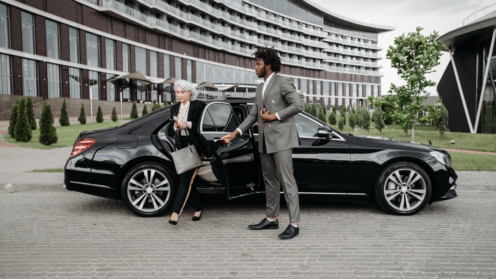 A professional chauffeur assists a businesswoman exiting a luxury car in front of a modern building.