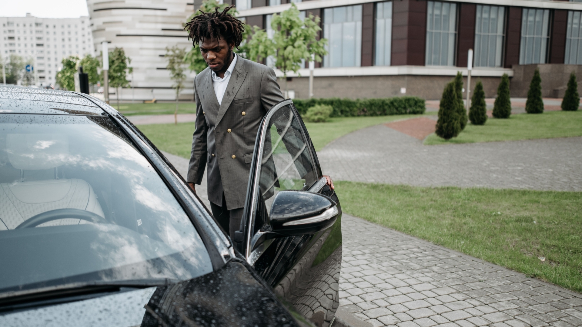 Man in a gray suit opening a black car door in an urban setting.